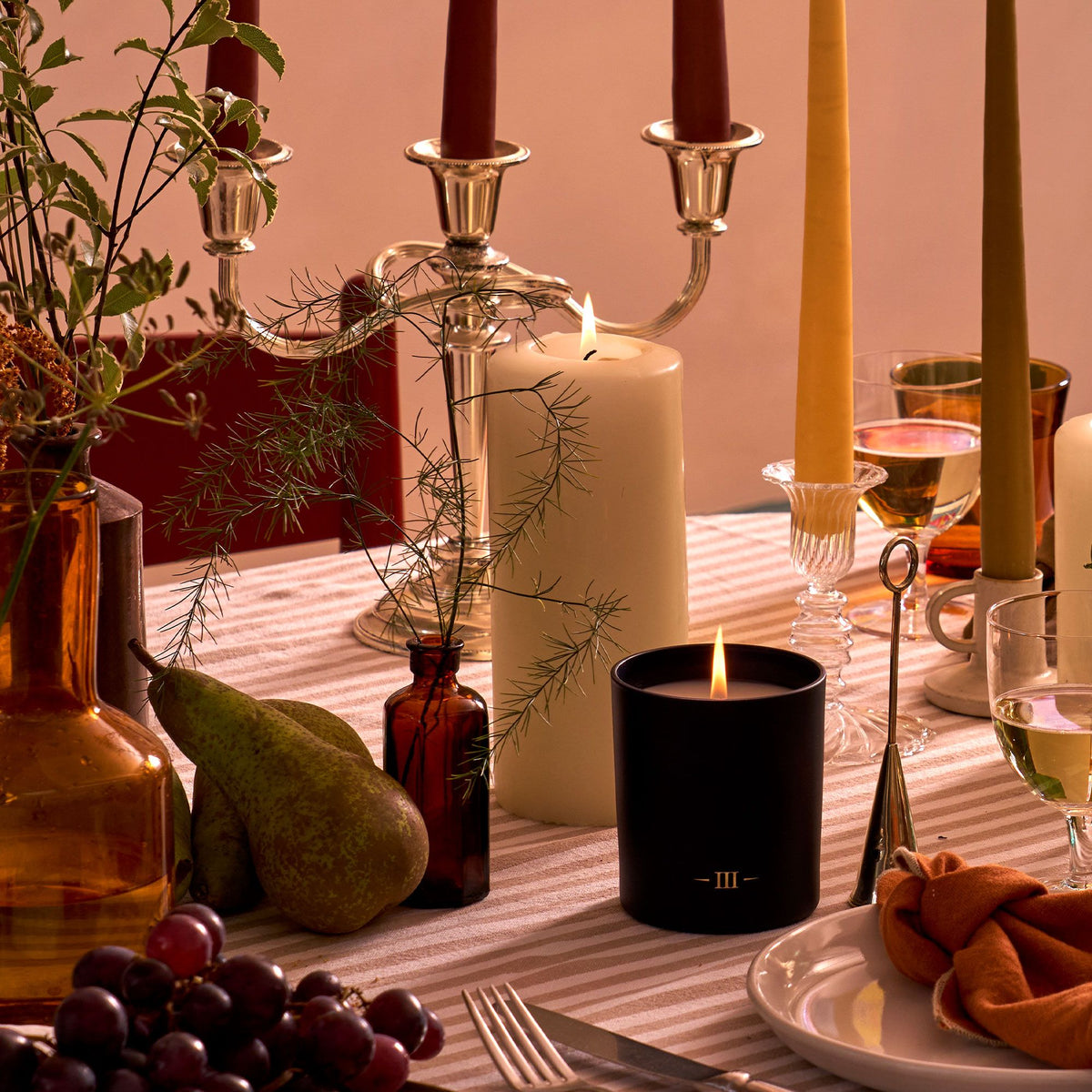 Charles Farris Rubus scented candle lit on an autumn dining table, with a cream pillar candle, burgundy, mustard and olive tapered candles in a silver candelabra, pears, grapes and amber glassware