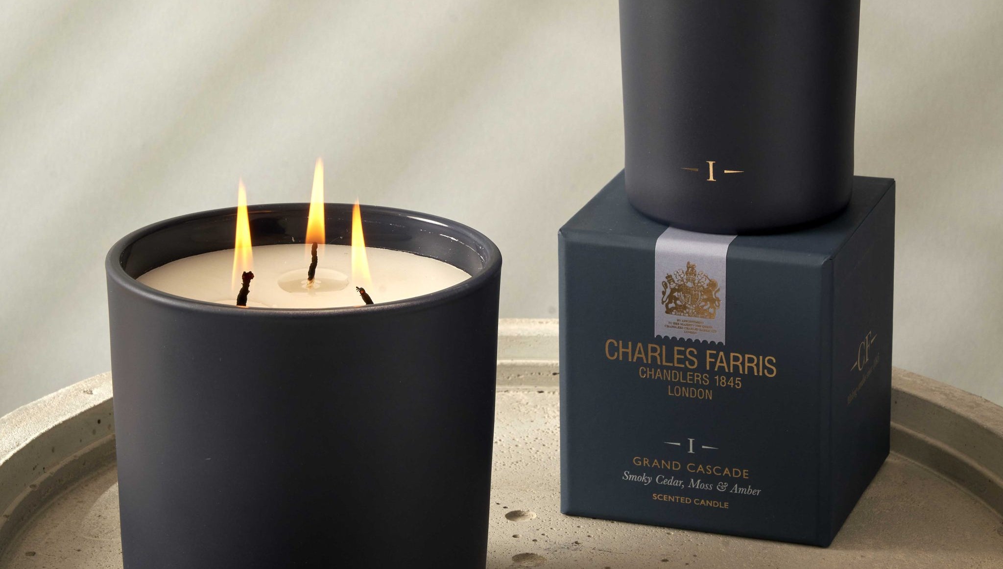 Two candles with lit wicks on a stone surface, one candle in front of its packaging box.
