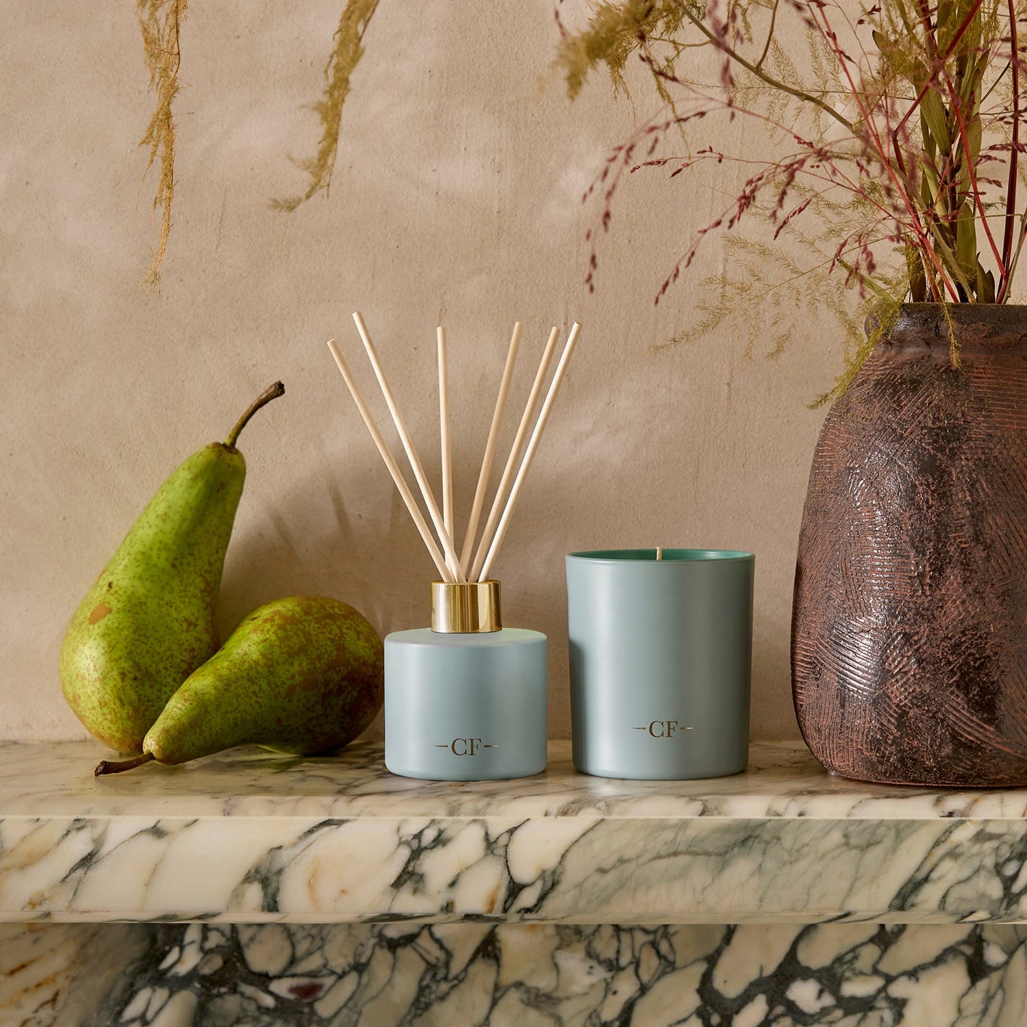 Decorative items including a diffuser, candle, and pears on a marble surface with a beige wall in the background.