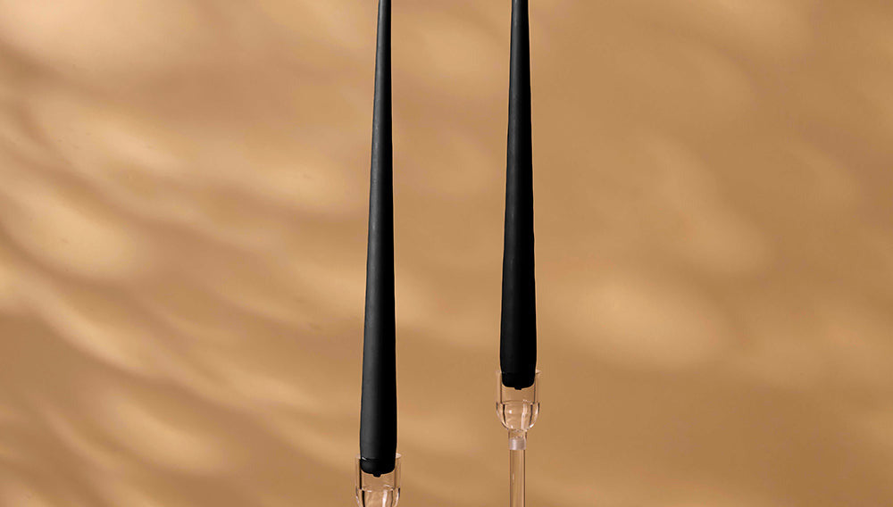 Two black candles on clear candlesticks against a beige wall.