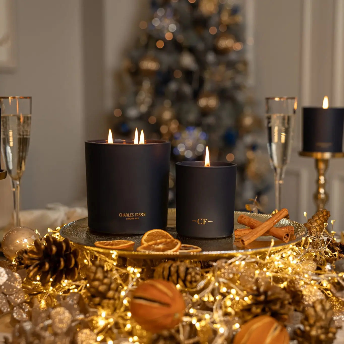 Two scented candles on a table with Christmas decorations including a tree, glasses, and pinecones.