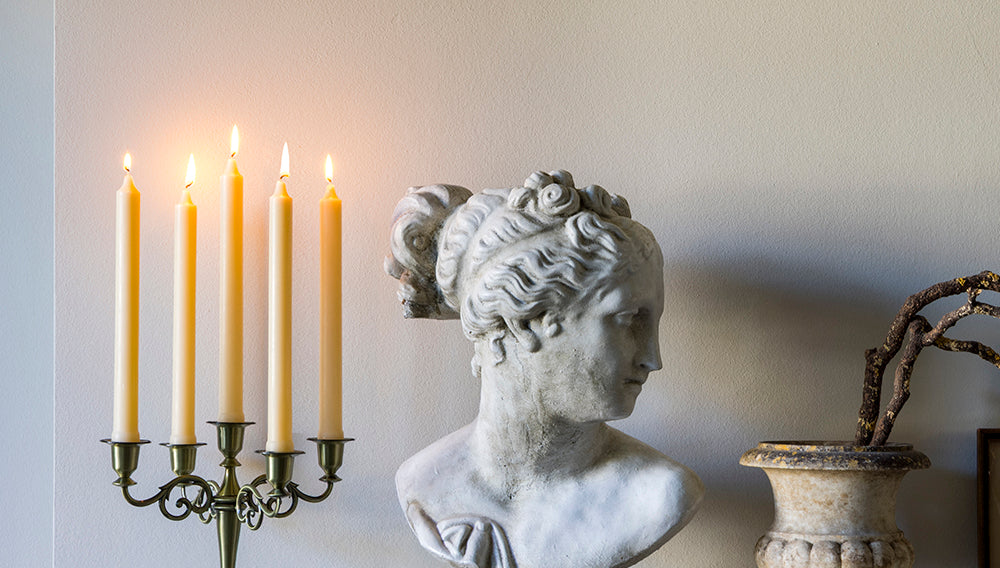 Bust of a classical figure, candle holder with lit candles, and decorative urn on a marble surface.
