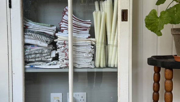 White cabinet with glass doors displaying folded clothes and bottles, as well as Charles Farris dining candles in glass containers.