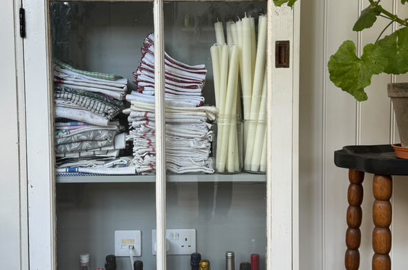 White cabinet with glass doors displaying folded clothes and bottles, as well as Charles Farris dining candles in glass containers.