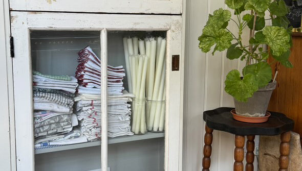 White cabinet with glass doors displaying folded clothes and bottles, as well as Charles Farris dining candles in glass containers.