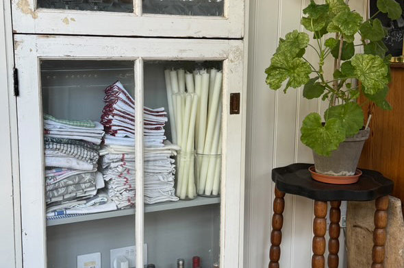 White cabinet with glass doors displaying folded clothes and bottles, as well as Charles Farris dining candles in glass containers.