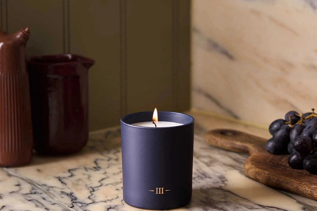 Candle in a dark blue holder on a marble surface with grapes and ceramic items in the background.