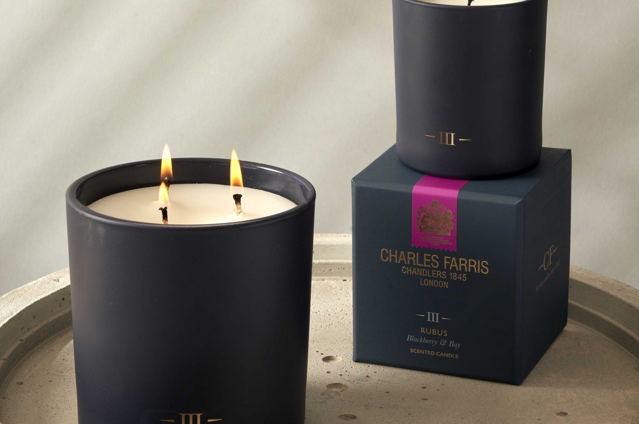 Two candles with visible flames on a stone surface, one on a box labeled 'Charles Farris'.