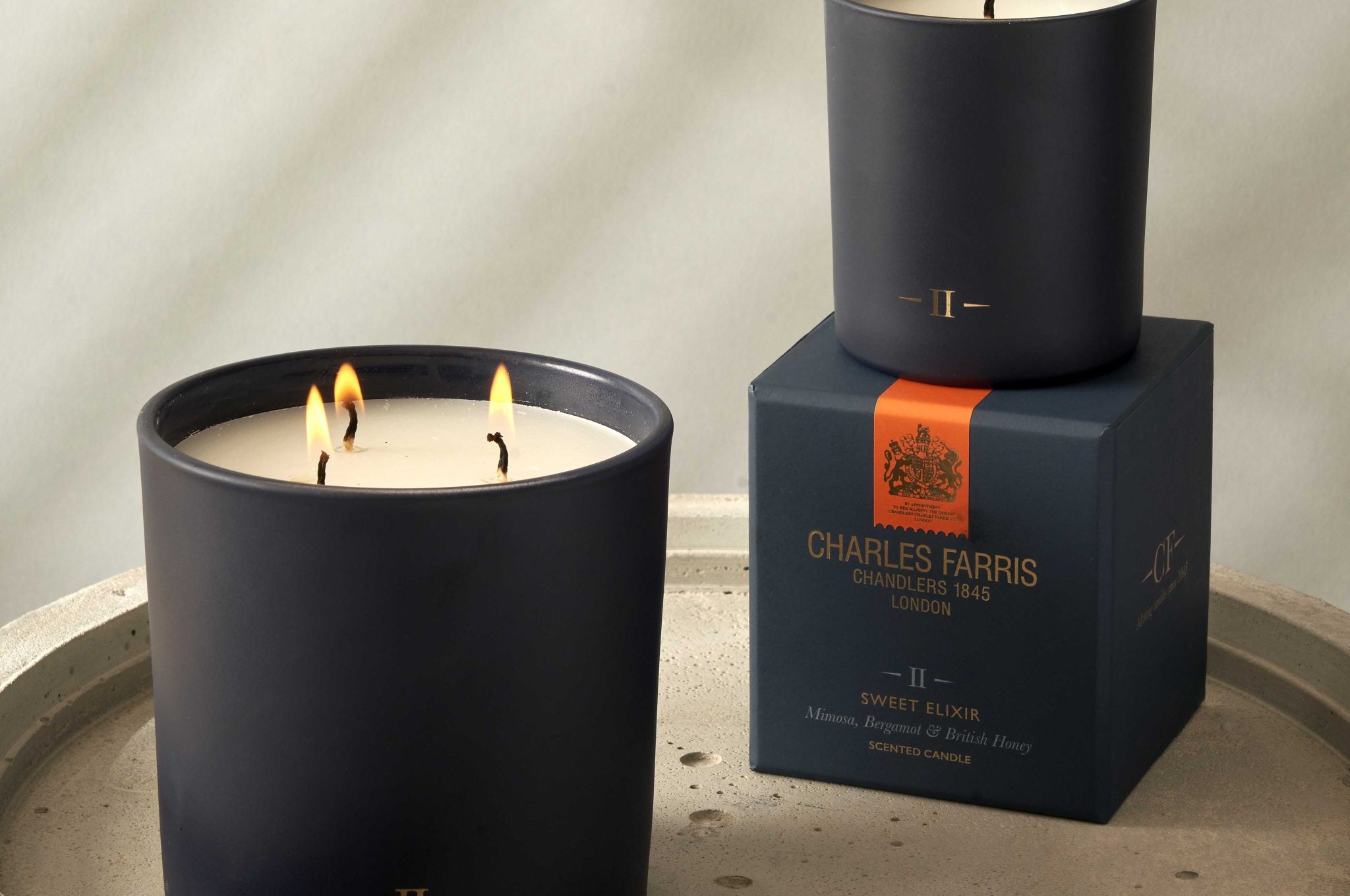 Two candles with visible flames on a stone surface, one on top of its packaging box featuring 'Charles Farris' branding.