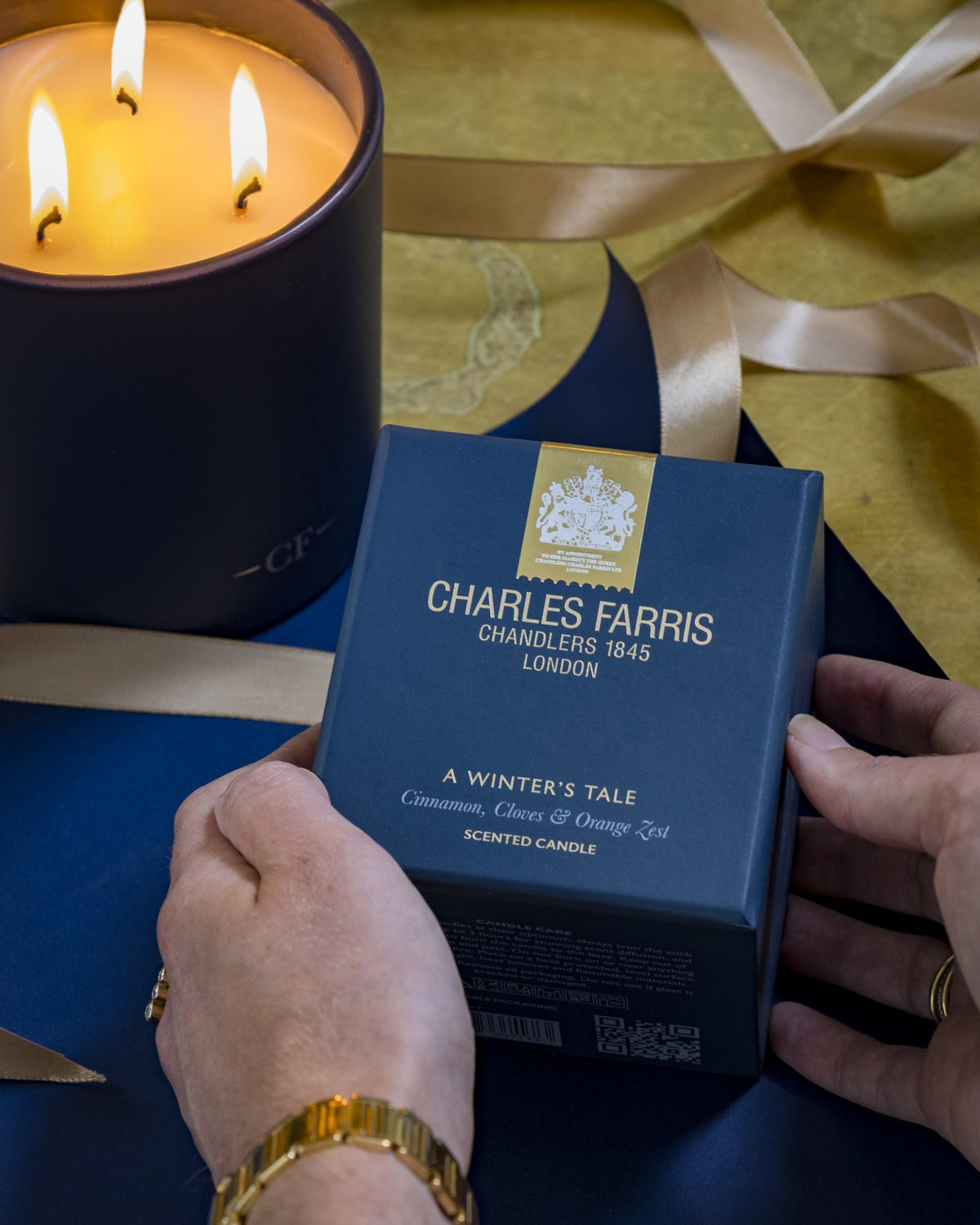 Hands holding a Charles Farris A Winter's Tale scented candle gift box beside a lit three-wick candle
