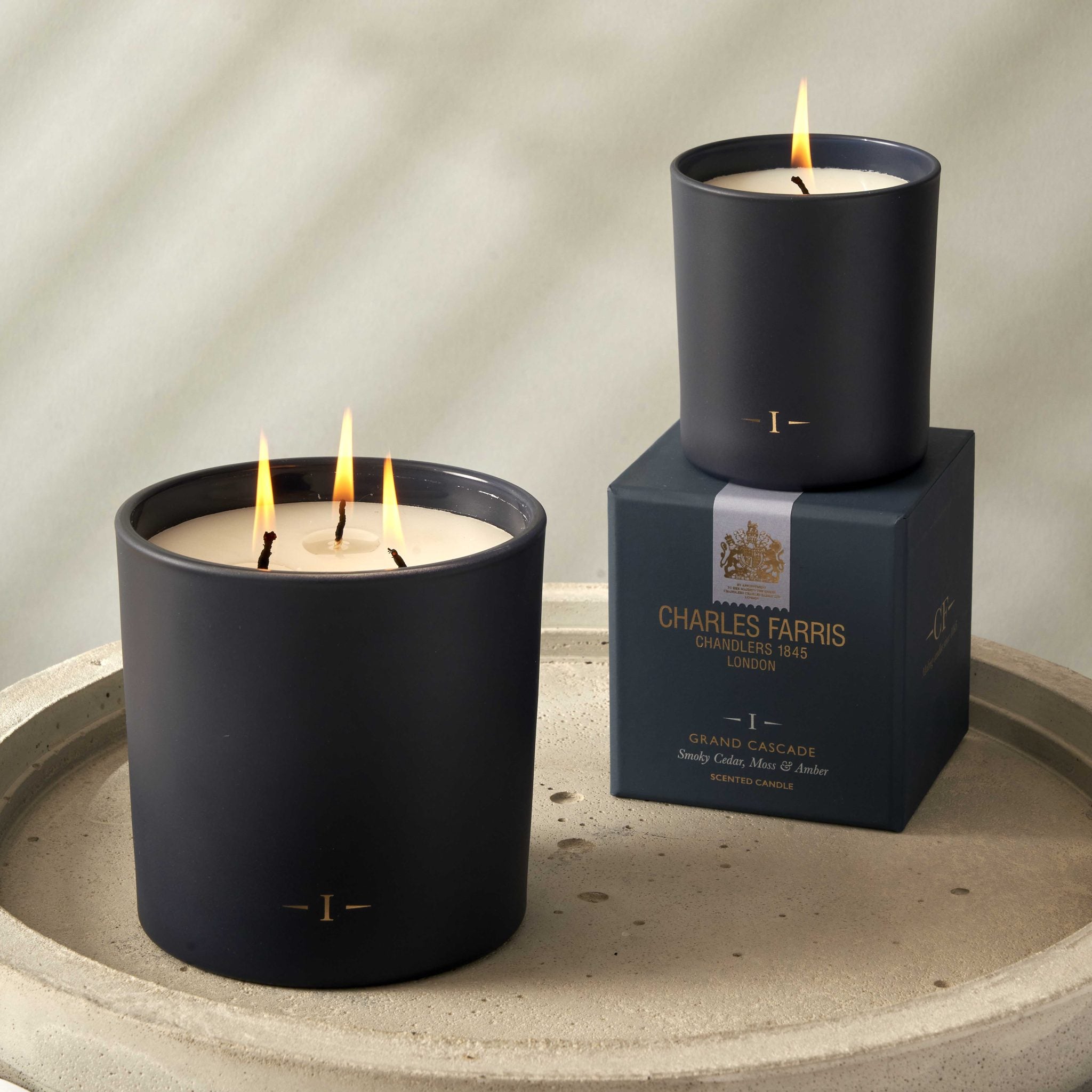 Two candles with lit wicks on a stone surface, one candle in front of its packaging box.