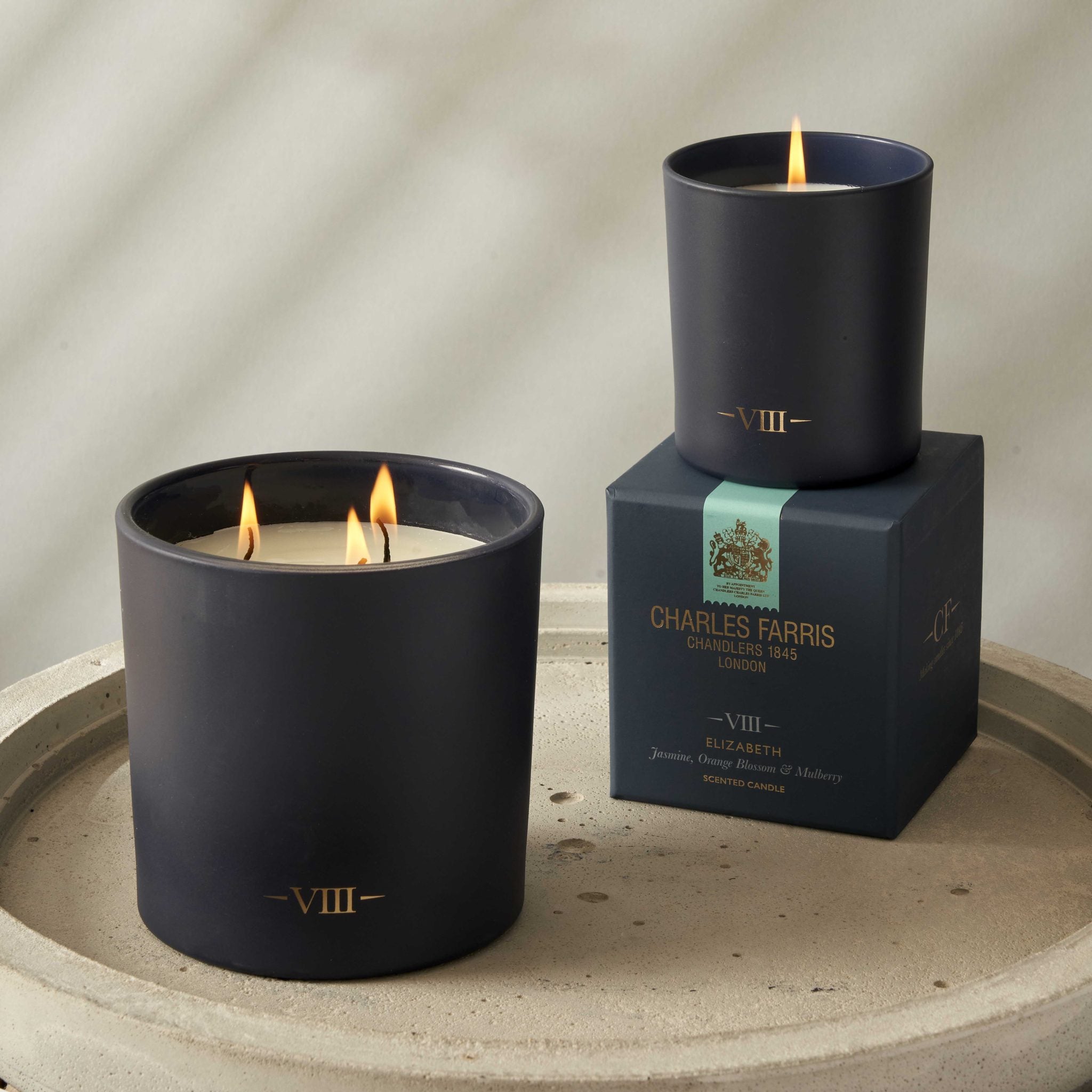 Two lit candles in black containers with a box labeled 'Charles Farris' on a neutral background.