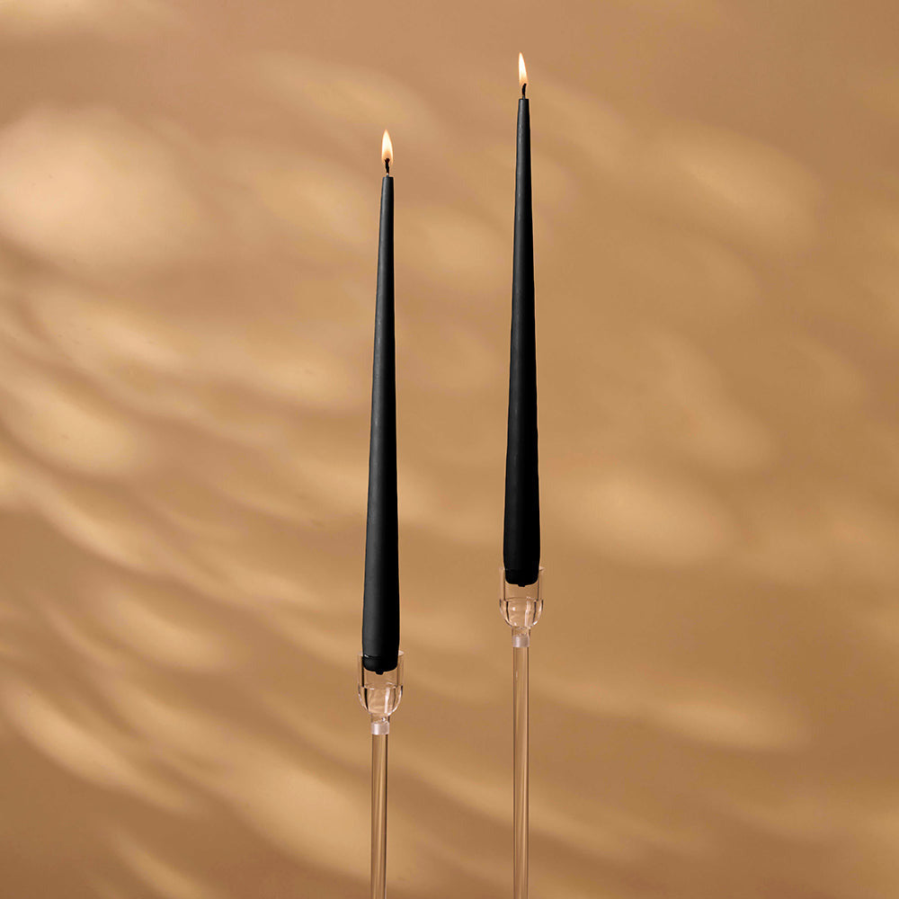 Two black candles on clear candlesticks against a beige wall.