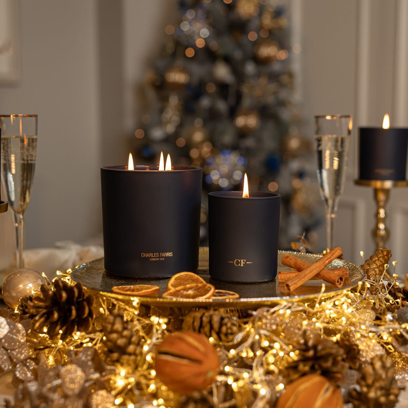 Two lit candles on a table with festive decorations and a blurred Christmas tree in the background.