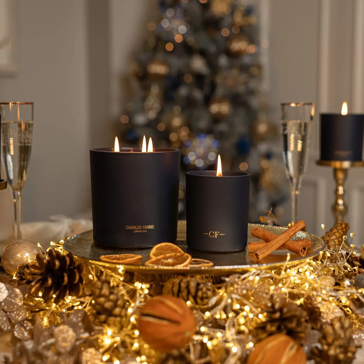 Two scented candles on a table with Christmas decorations including a tree, glasses, and pinecones.