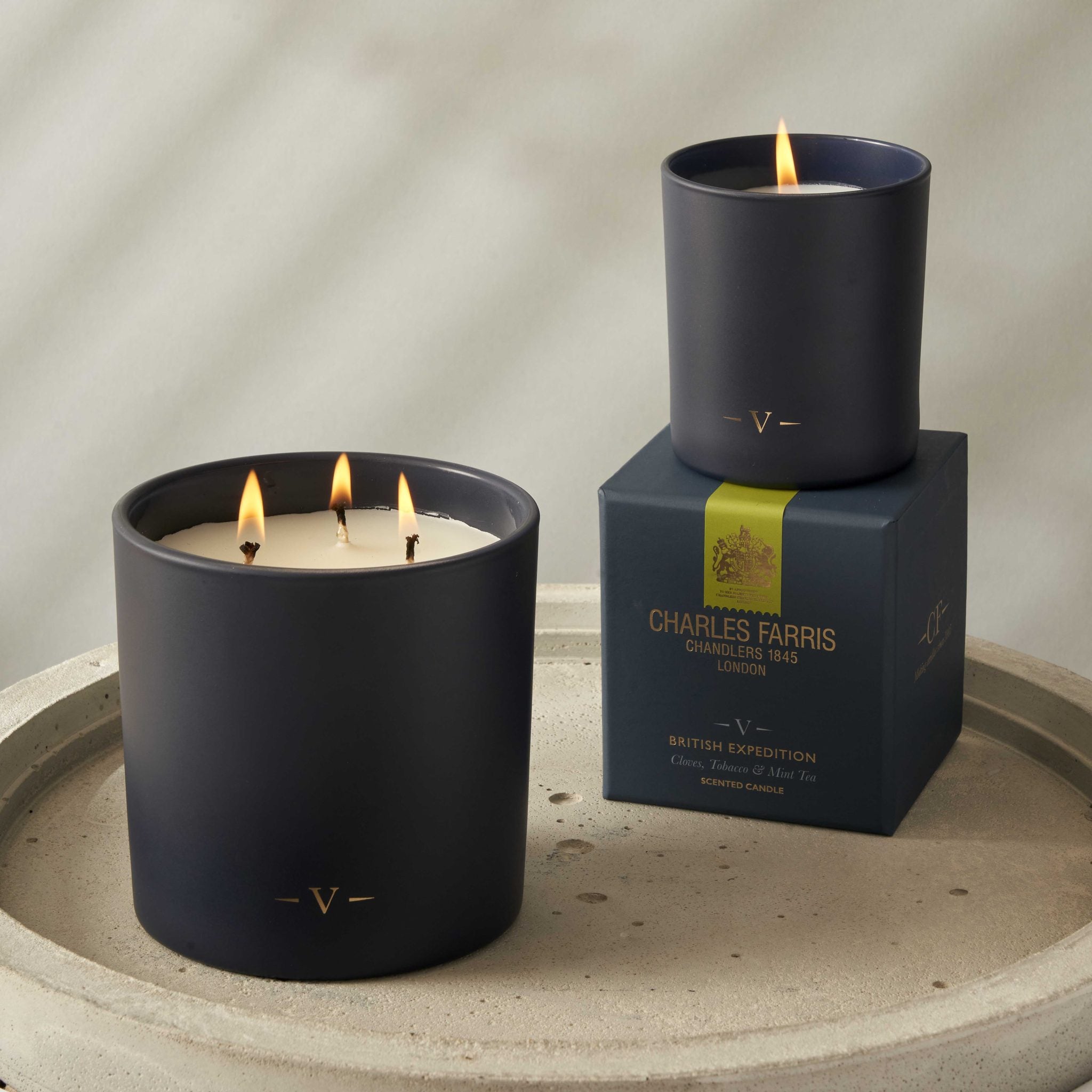 Two candles with visible flames on a stone surface, one candle on top of a branded box.