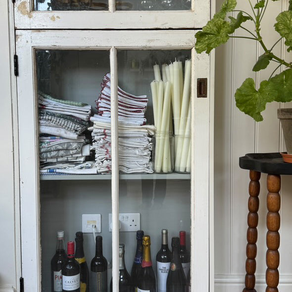 White cabinet with glass doors displaying folded clothes and bottles, as well as Charles Farris dining candles in glass containers.