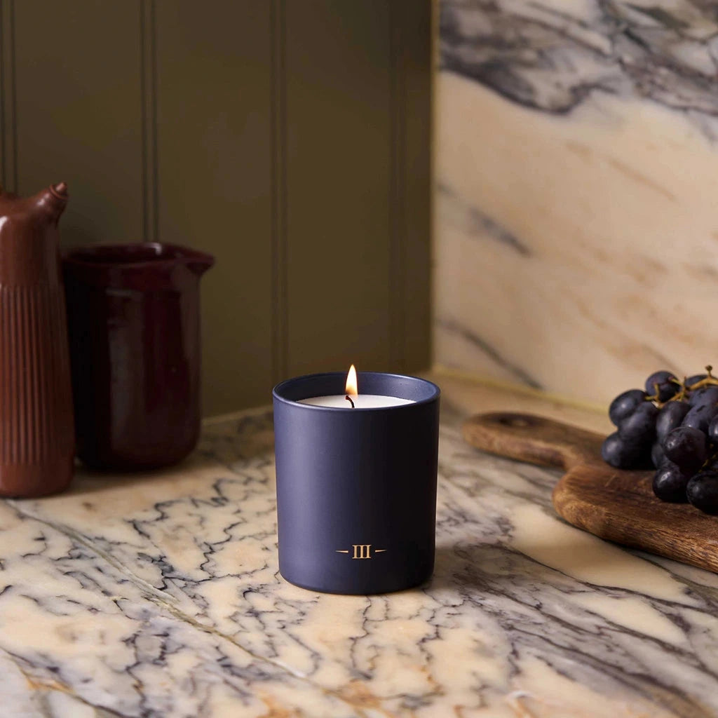 Candle in a dark blue holder on a marble surface with grapes and ceramic items in the background.