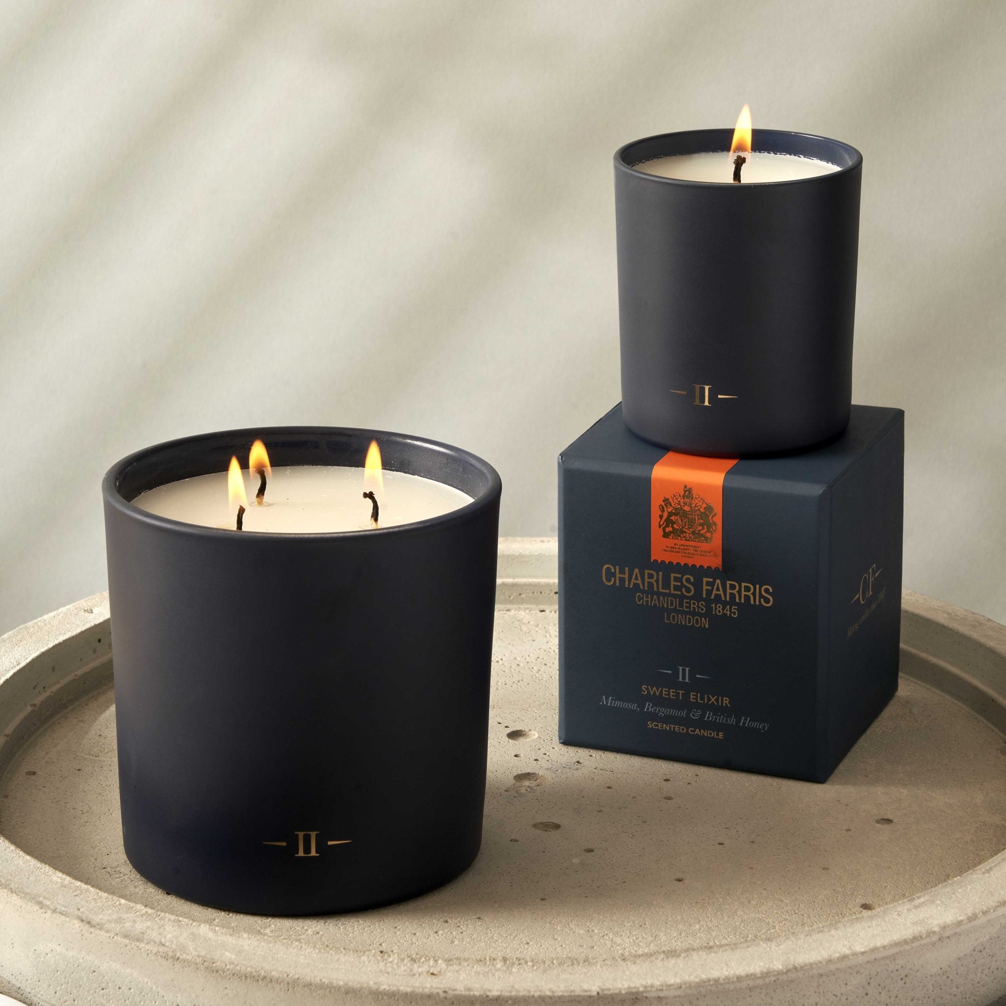 Two candles with visible flames on a stone surface, one on top of its packaging box featuring 'Charles Farris' branding.