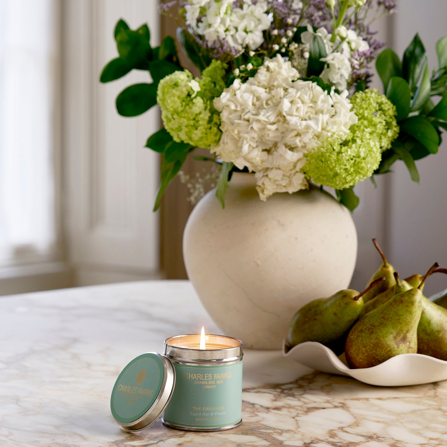 The Orchard Tin Candle on a marble surface with a vase of flowers