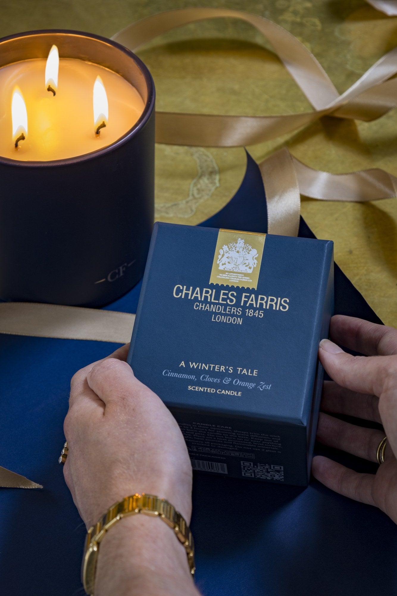 Hands holding a Charles Farris A Winter's Tale scented candle gift box beside a lit three-wick candle