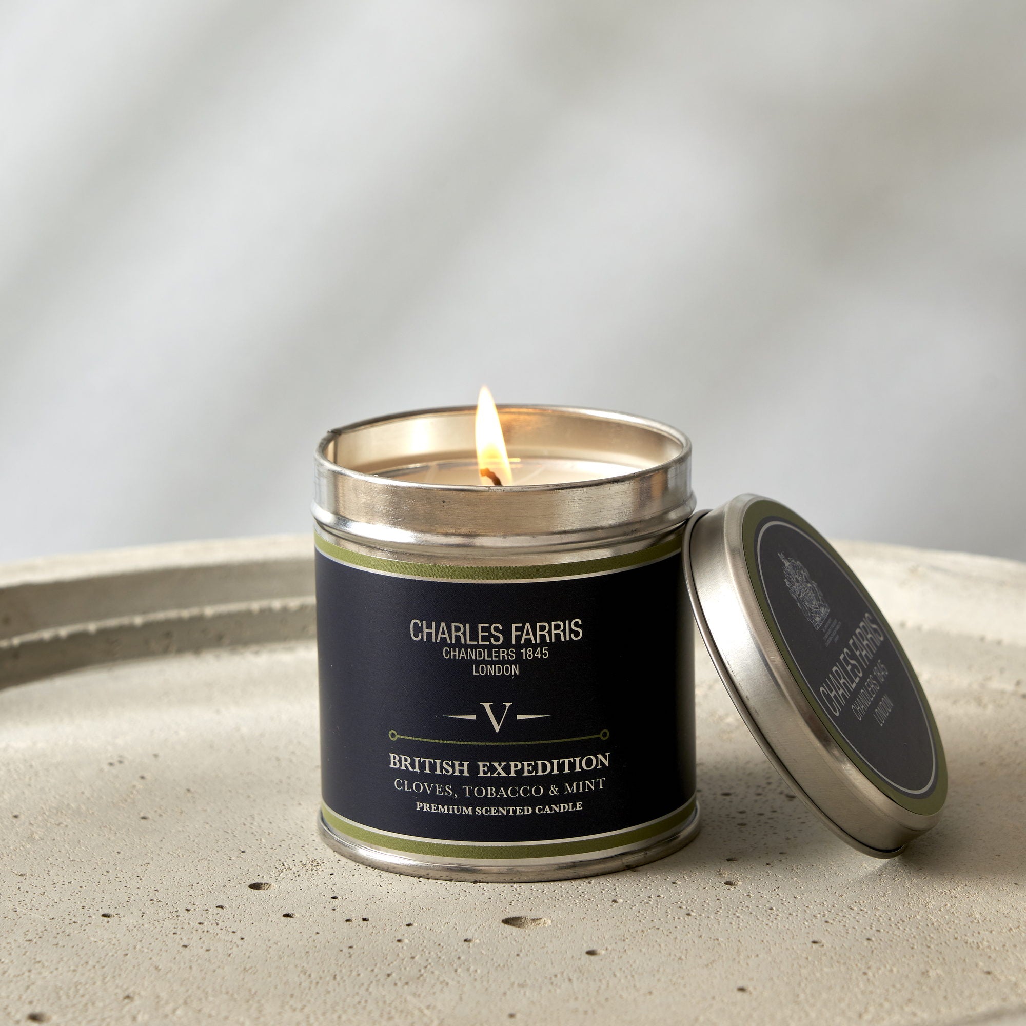 Charles Farris British Expedition signature tin candle lit with its lid resting alongside, cloves, tobacco and mint scent