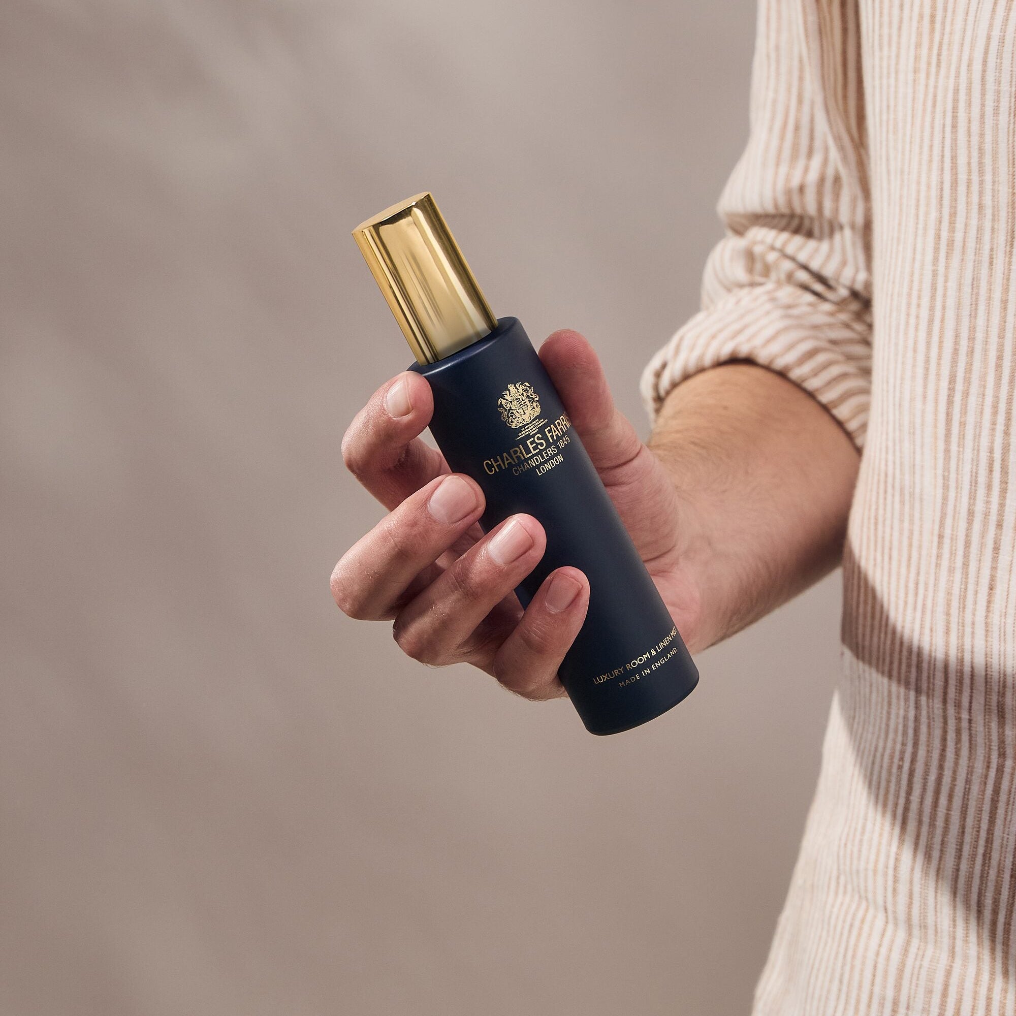 Person holding a dark navy blue bottle of Charles Farris room spray scented home mist against a neutral beige background.