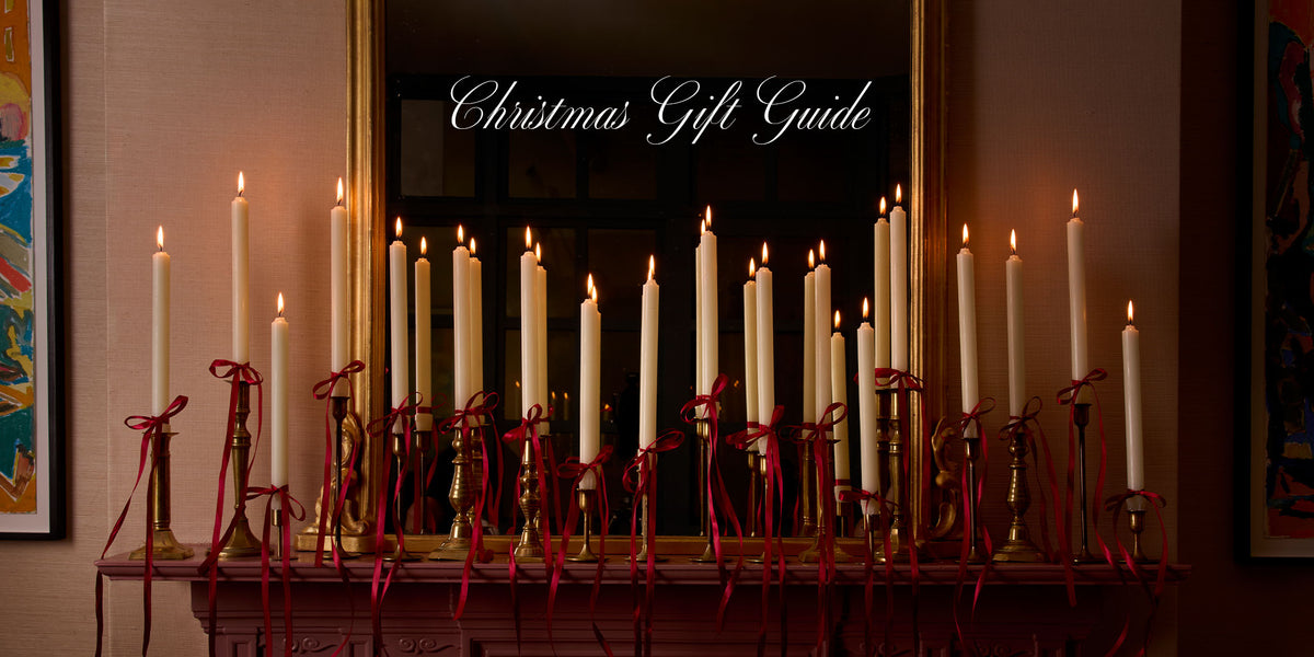 Christmas Gift Guide: Charles Farris ivory dinner candles tied with red ribbon, lit along a mantelpiece